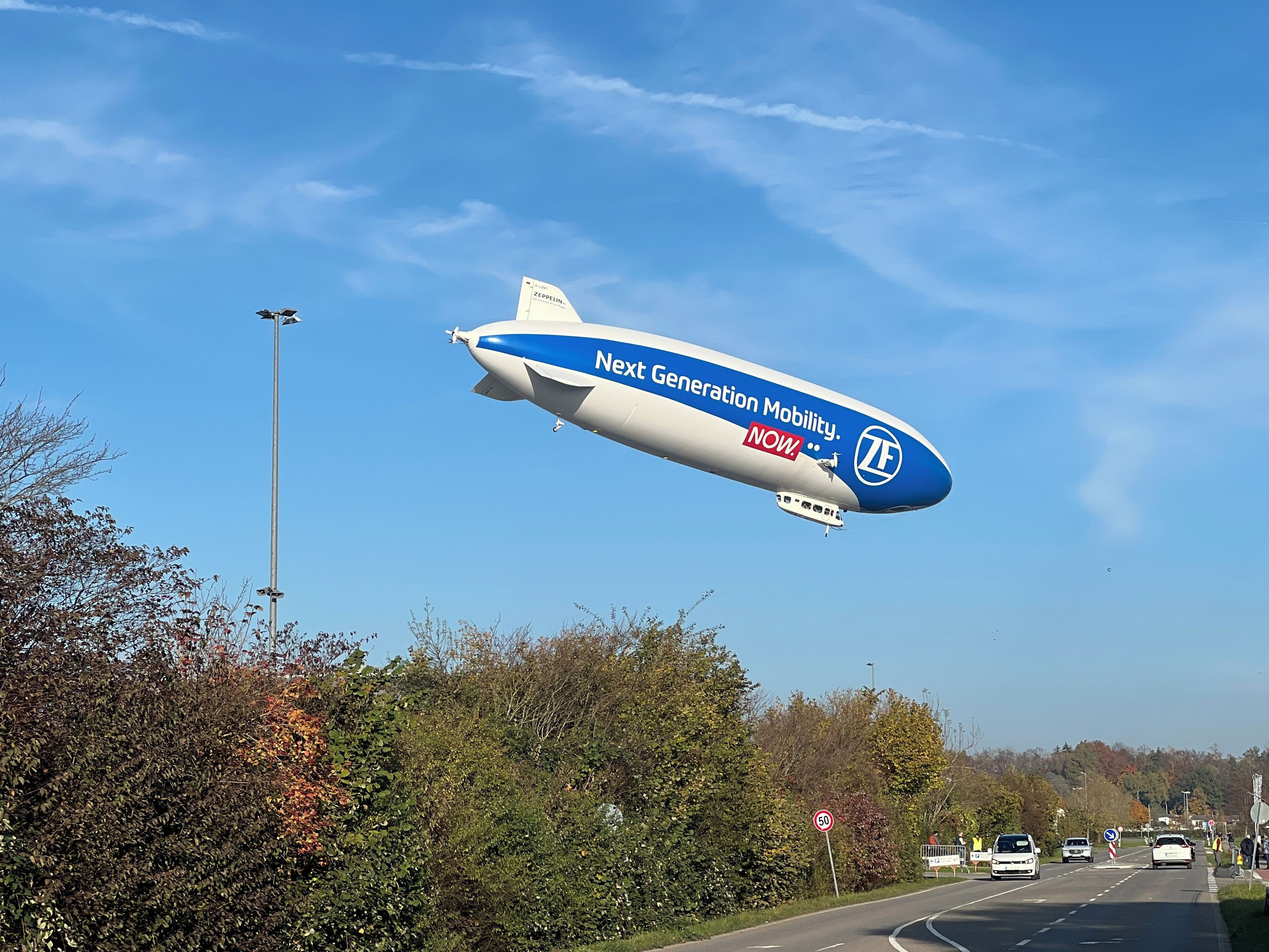 A zeppelin flying over a road with autumn trees and a speed limit sign.