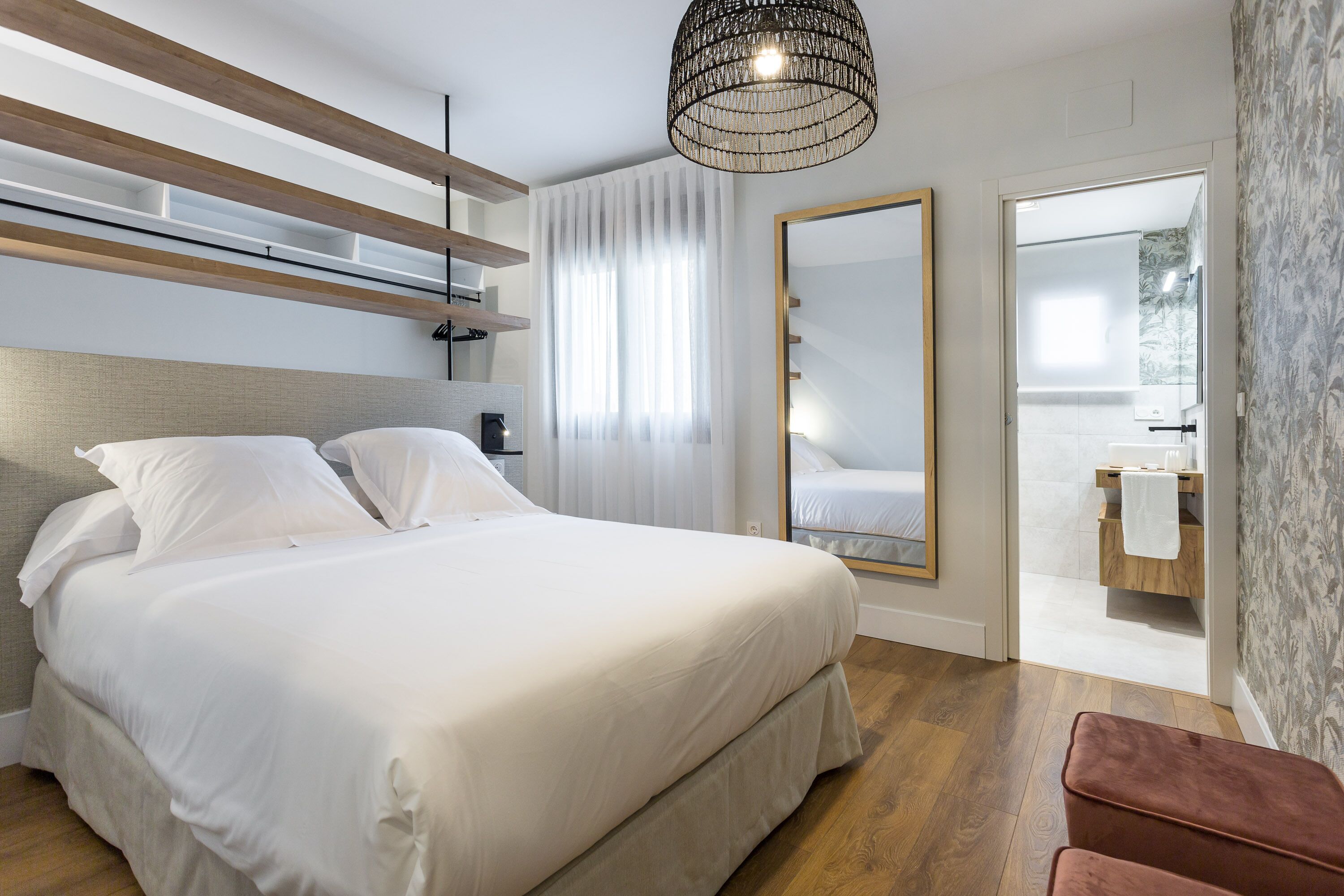 Un dormitorio moderno con una cama grande, vigas de madera, una lámpara colgante y un baño visible a través de una puerta abierta.