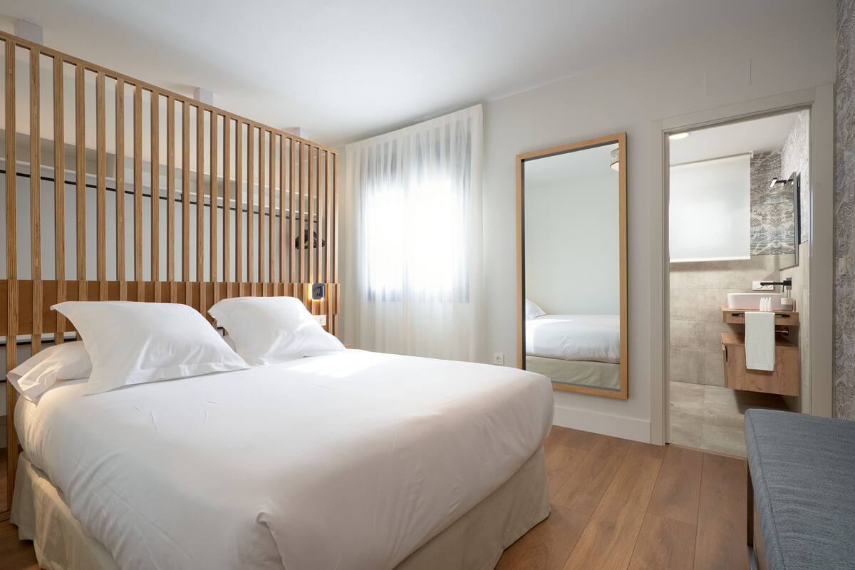 Quarto de hotel moderno com uma cama grande, cabeceira de madeira, um espelho, banheiro com pia e toalha, e piso de madeira.