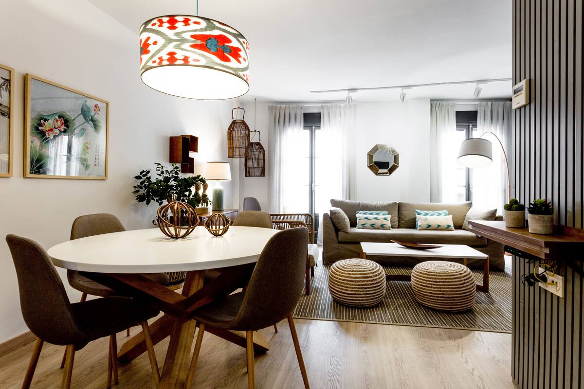 Uma sala de estar moderna com uma mesa de jantar redonda, cadeiras marrons, um sofá com almofadas, uma mesa de centro e um lustre.