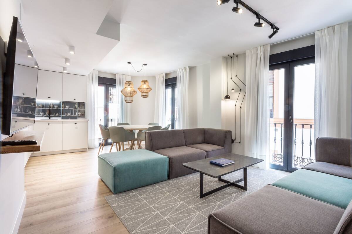 Uma sala de estar moderna com sofá, banquinho e mesa de centro. Há uma área de cozinha com armários e uma mesa de jantar. O quarto possui grandes janelas com cortinas e uma varanda com guarda-corpo.