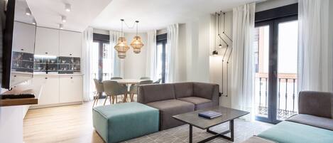 A modern living room with a sofa, ottoman, and coffee table. There is a kitchen area with cabinets and a dining table. The room has large windows with curtains and a balcony with a railing.