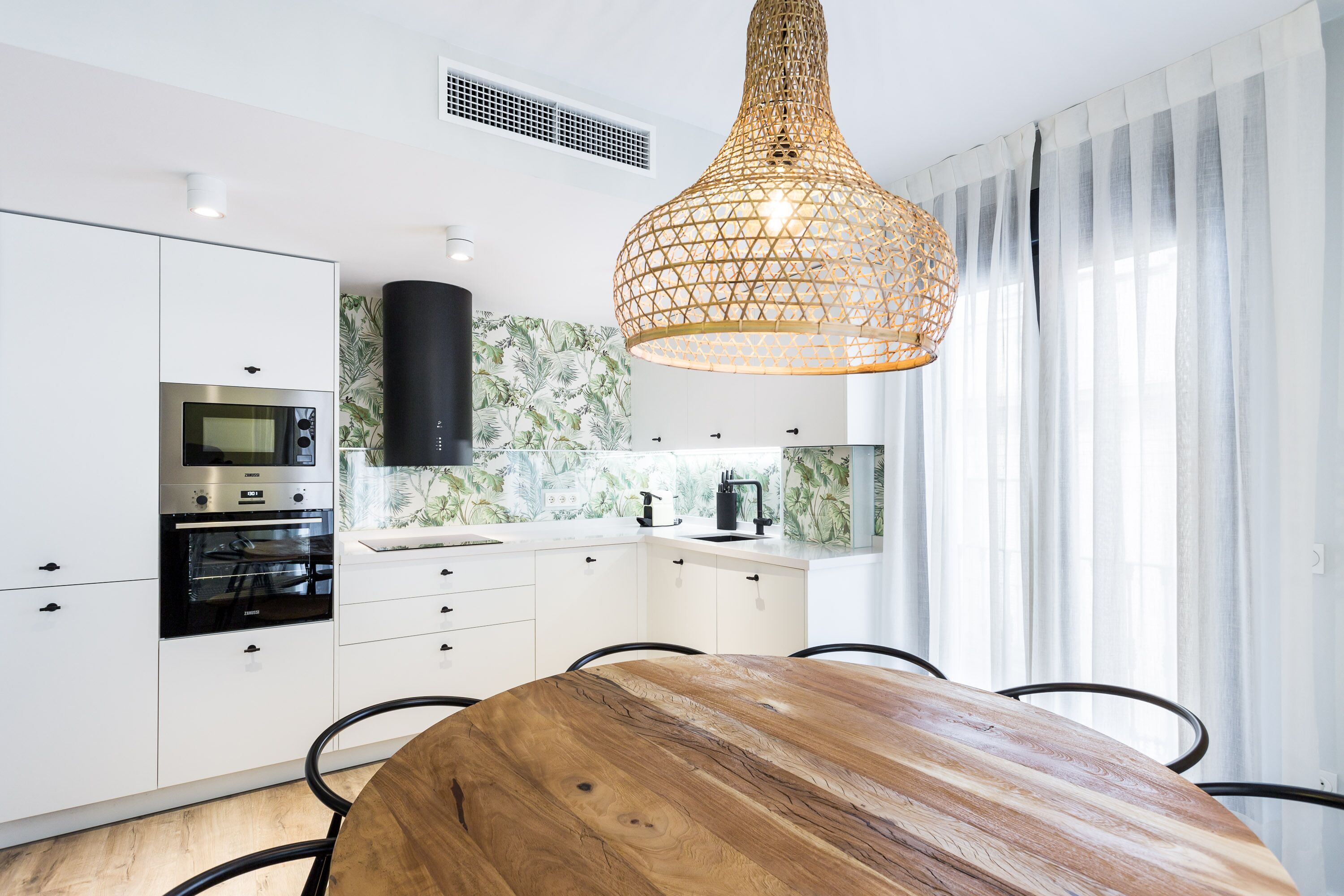 A modern kitchen with white cabinets, a wooden dining table, and a hanging light fixture.