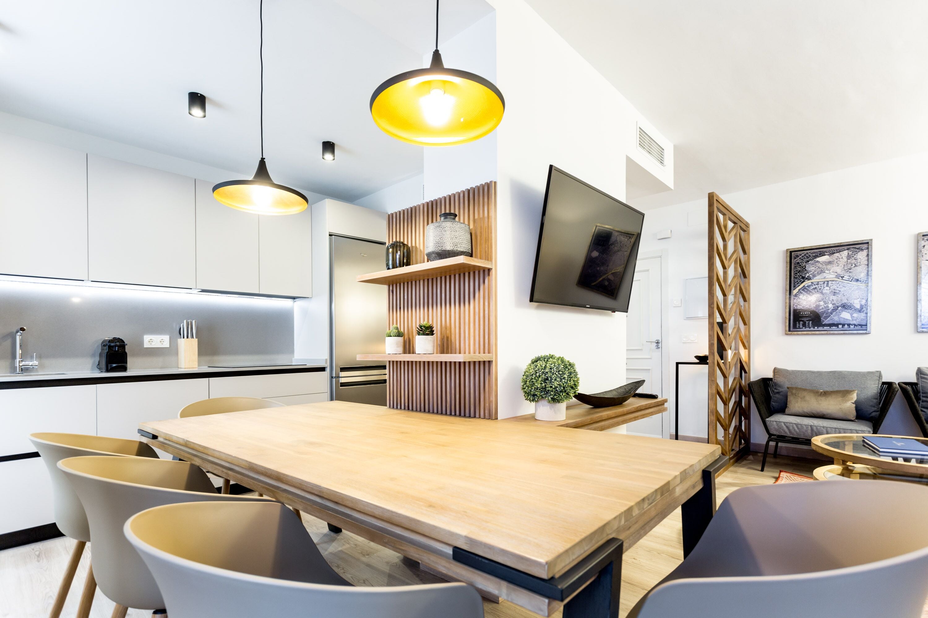 A modern kitchen with a wooden dining table, white chairs, pendant lights, and a built-in shelf with decorative items.