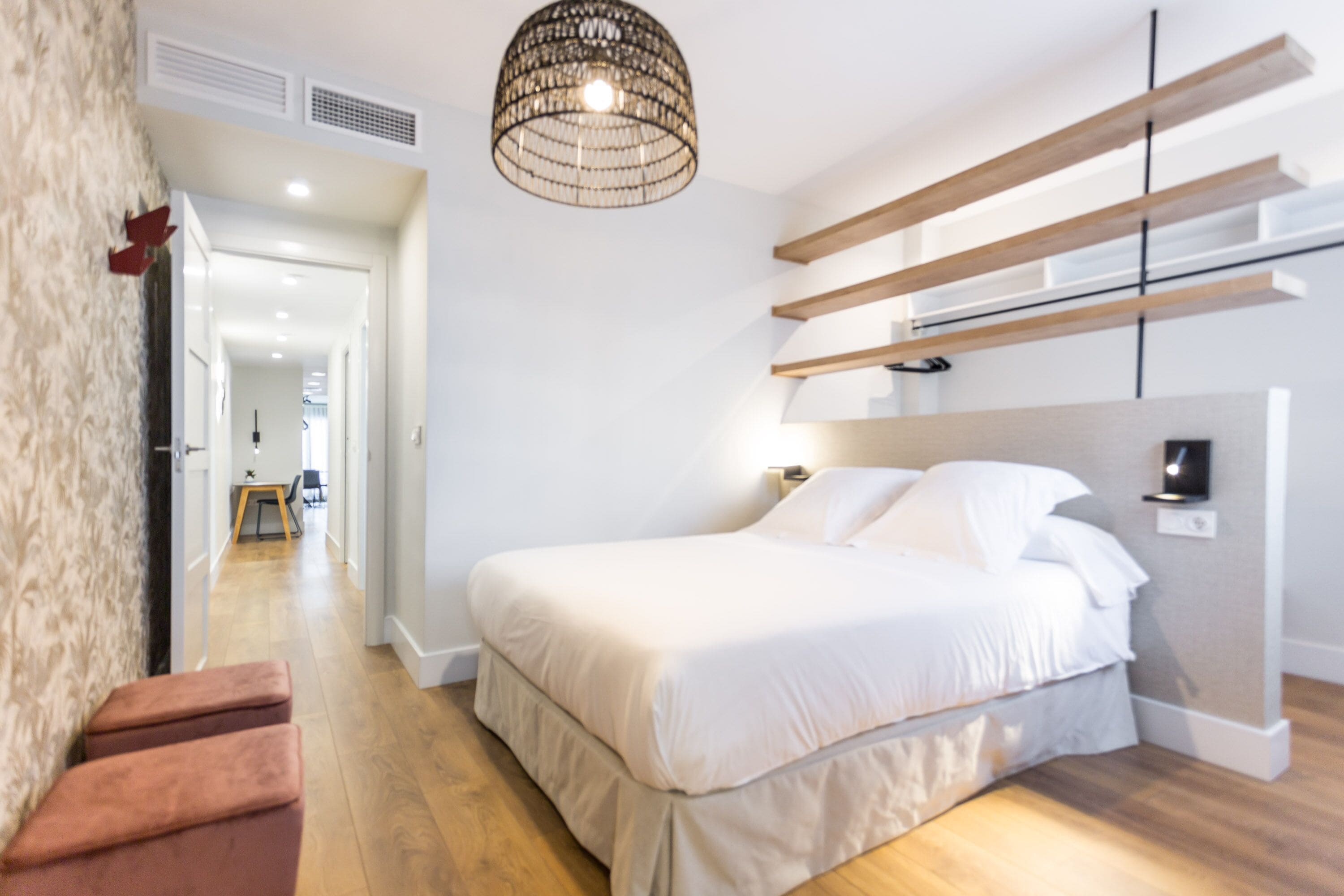 A modern bedroom with a large bed, wooden beams on the wall, a hanging light fixture, and a view into the adjacent room.