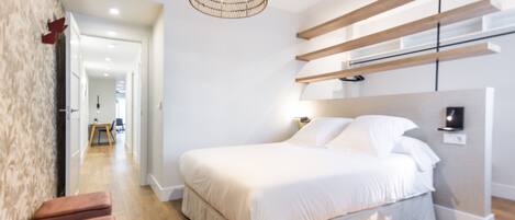 A modern bedroom with a large bed, wooden beams on the wall, a hanging light fixture, and a view into the adjacent room.