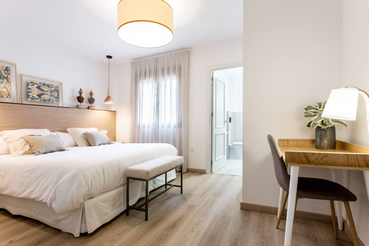 Quarto moderno com cama grande, mesa de cabeceira, escrivaninha com cadeira e janela com cortinas.