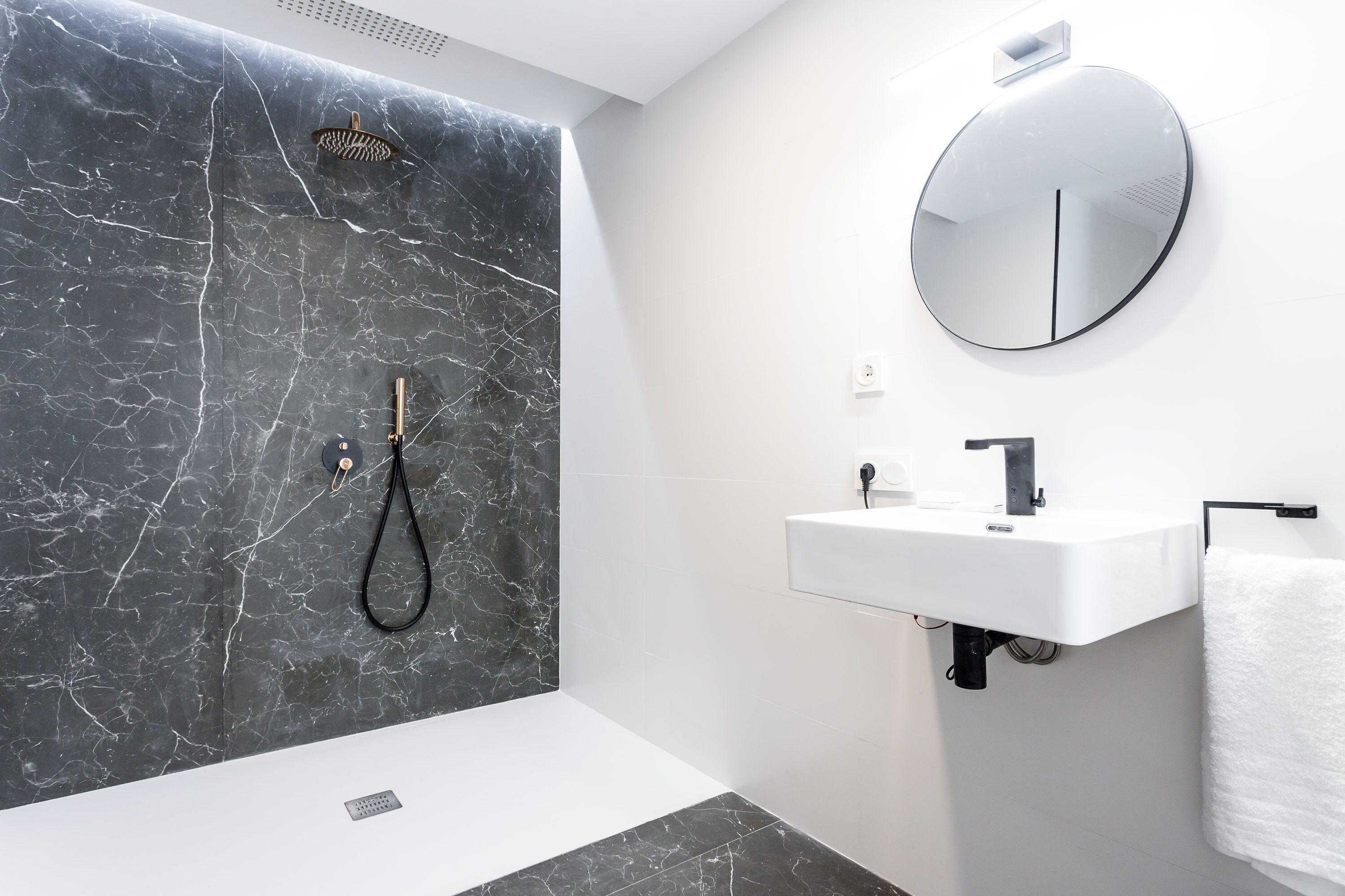 Une salle de bain moderne avec un mur de douche en marbre, un miroir rond, un lavabo blanc et un porte-serviettes.