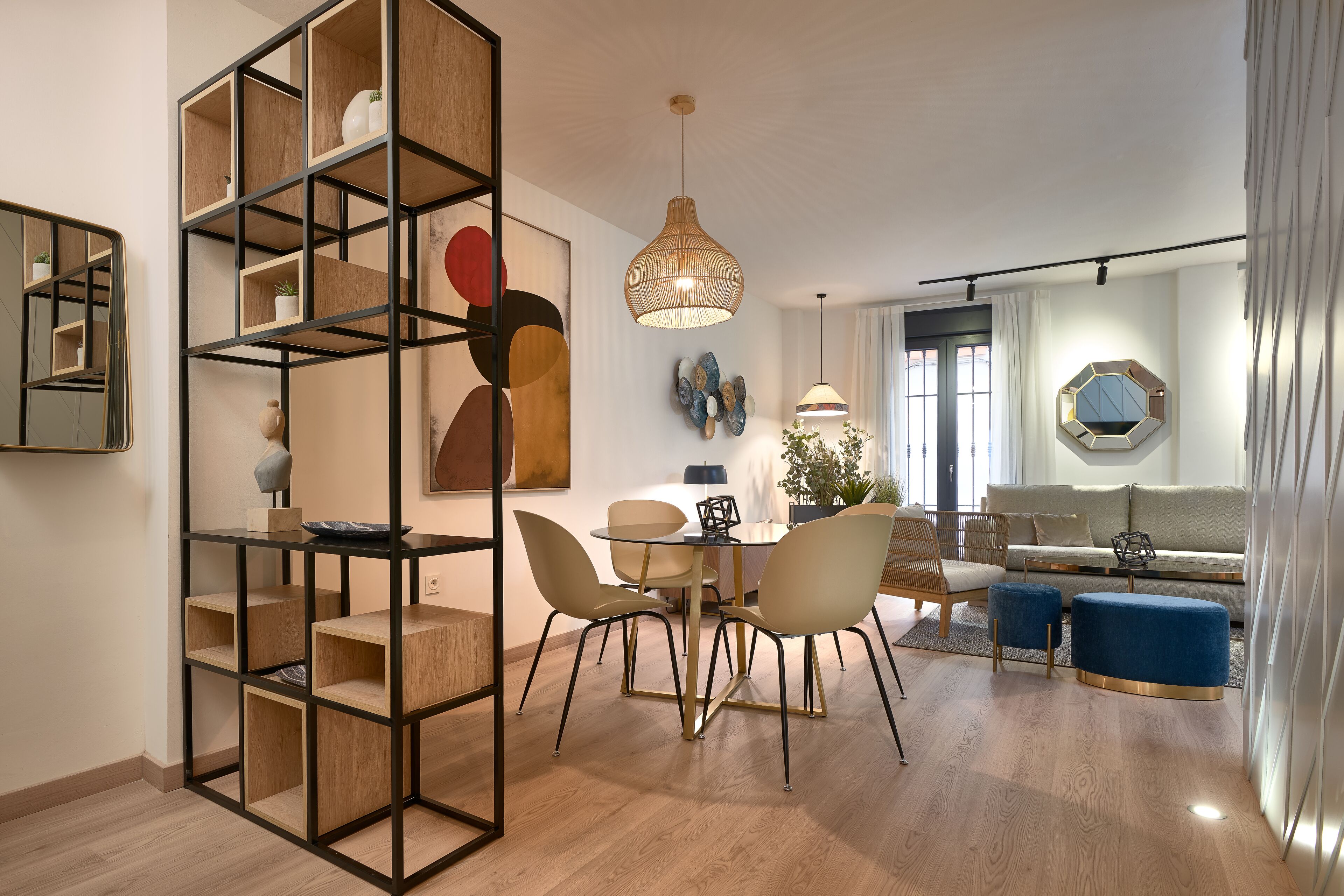 A modern living room with a dining area, featuring a wooden bookshelf, a round dining table with chairs, a sofa, and a large window with curtains.