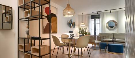 A modern living room with a dining area, featuring a wooden bookshelf, a round dining table with chairs, a sofa, and a large window with curtains.