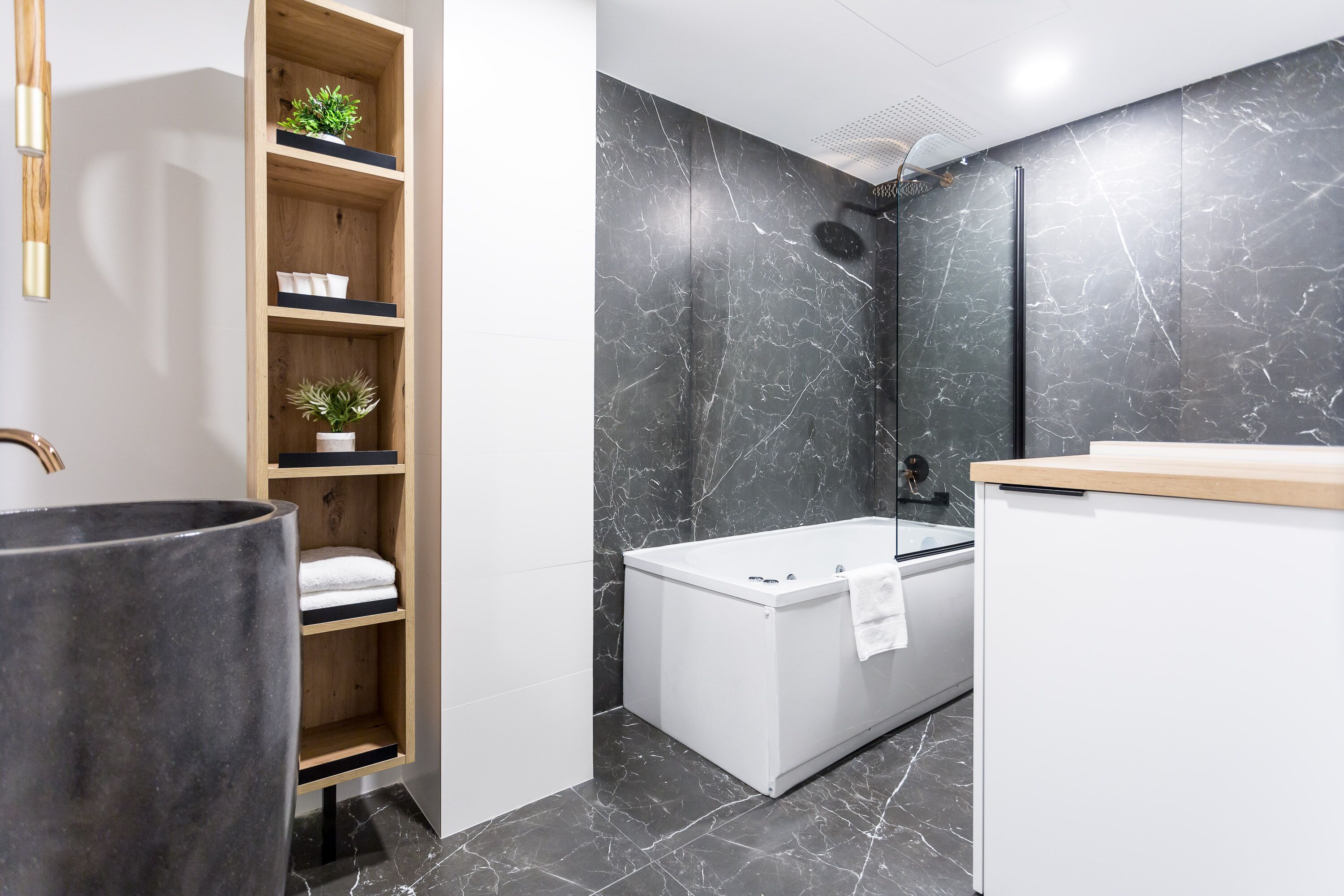 A modern bathroom with a white bathtub, a wooden shelf with plants, and a marble wall.