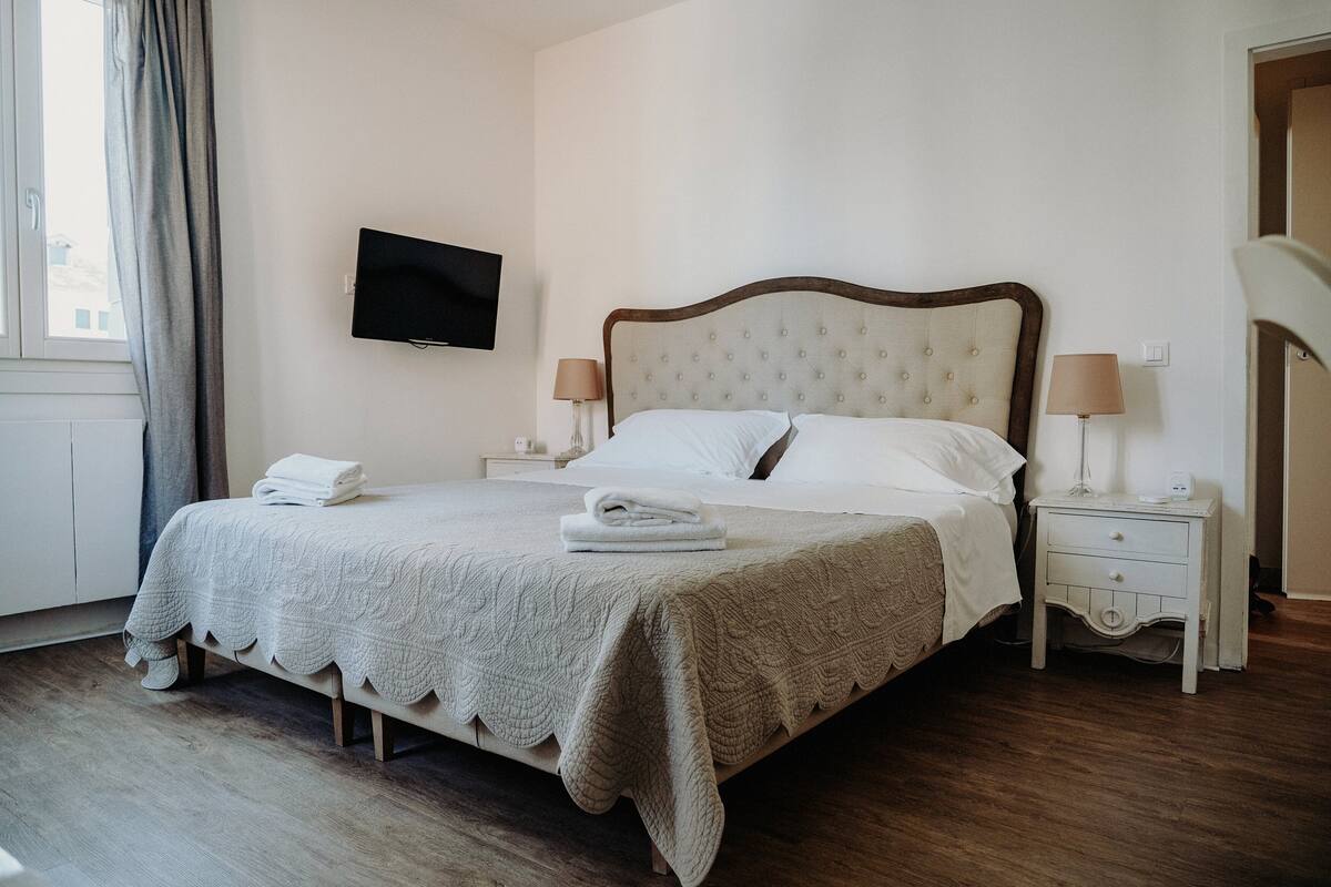 A neatly made bed with a quilted bedspread, two bedside tables with lamps, a flat-screen TV mounted on the wall, and a window with curtains.