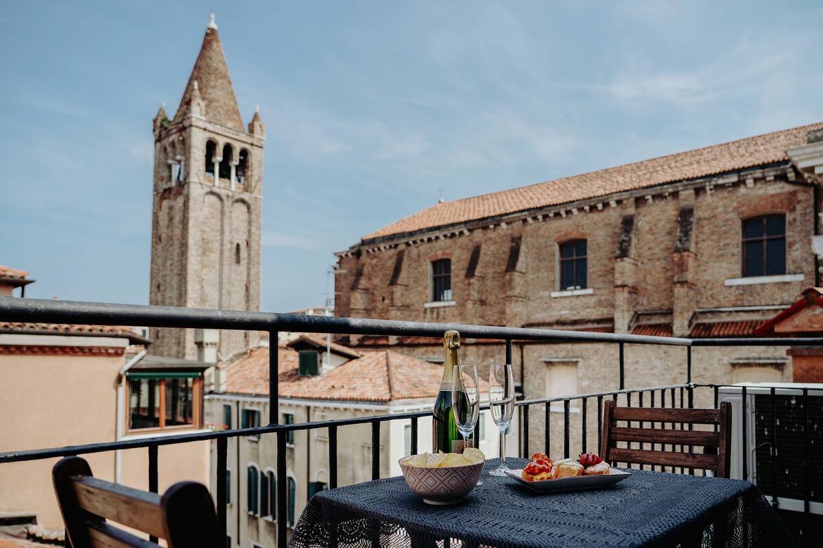 A balcony with a table set for a meal, a bottle of champagne, and a bowl of chips. In the background, there is a historic building with a tower.