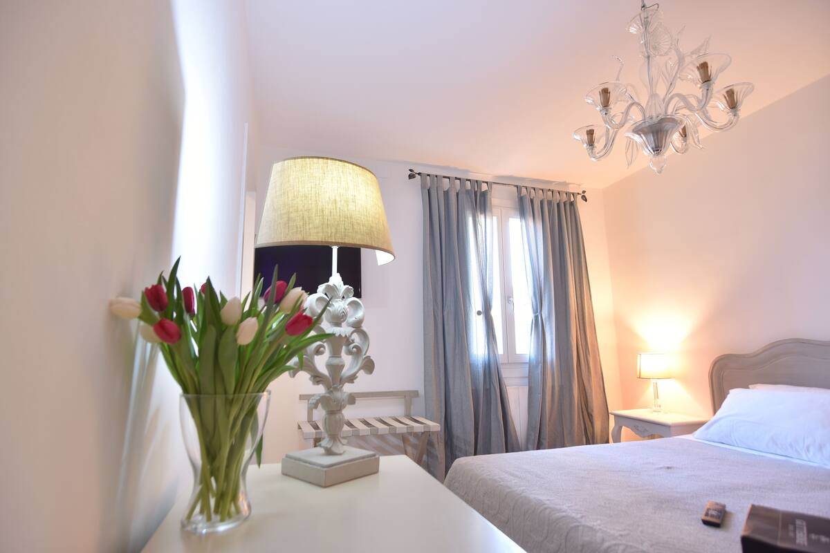 A hotel room with a bed, a bedside table with a lamp, a vase with flowers, and a chandelier.