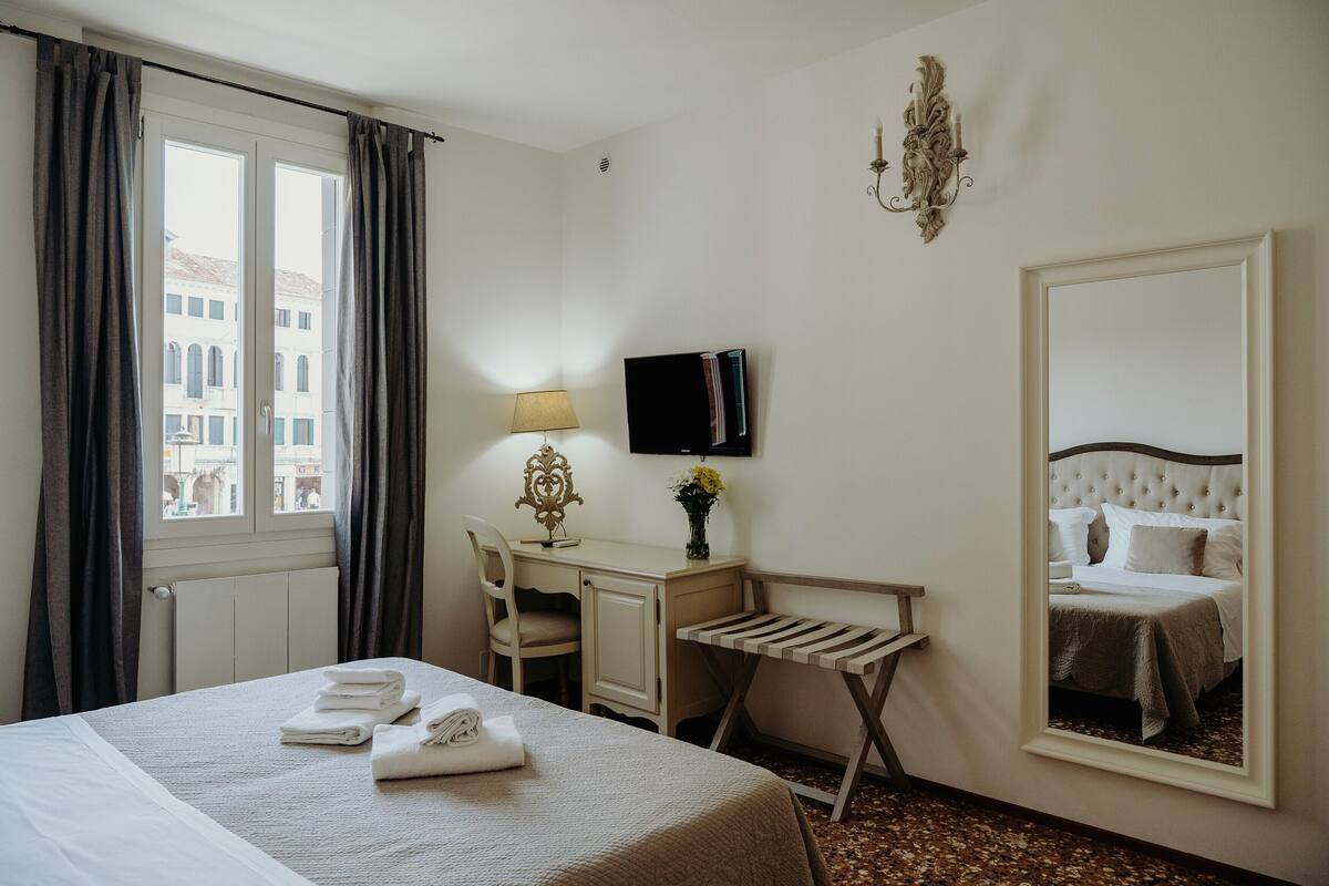 A hotel room with a bed, a desk, a chair, a small table, a mirror, and a window with curtains.