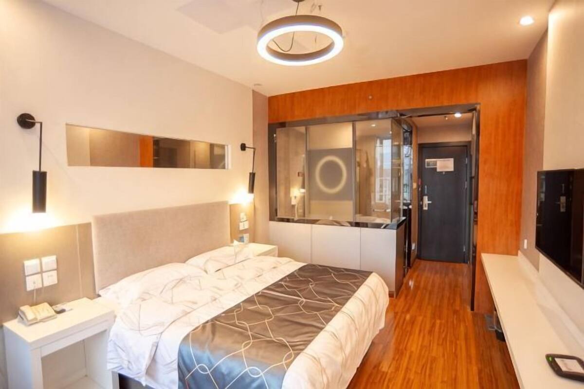 A modern hotel room with a large bed, bedside tables, a mirror, a wooden accent wall, and a door leading to a bathroom.