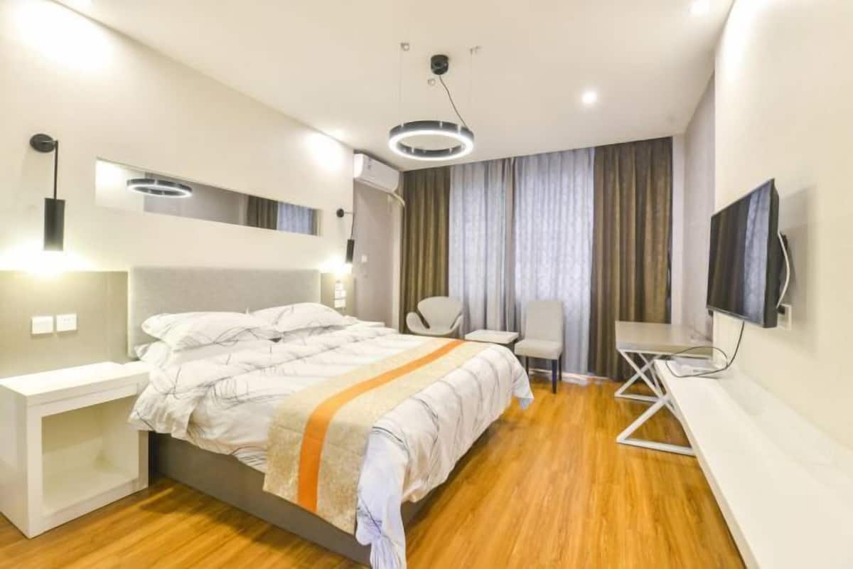 A modern hotel room with a bed, bedside table, chair, TV, and a ceiling light fixture.