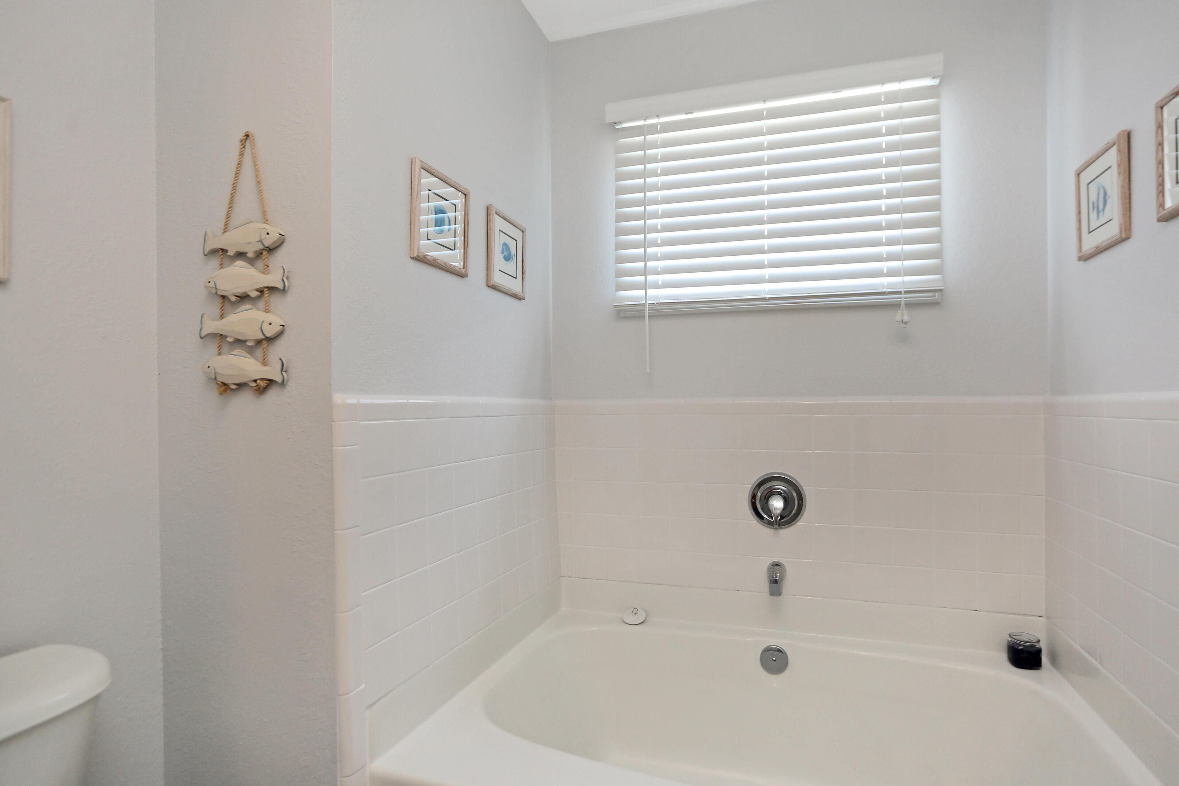 Un bagno con una vasca, pareti piastrellate di bianco, una finestra con persiane e una decorazione a forma di pesce appesa al muro.
