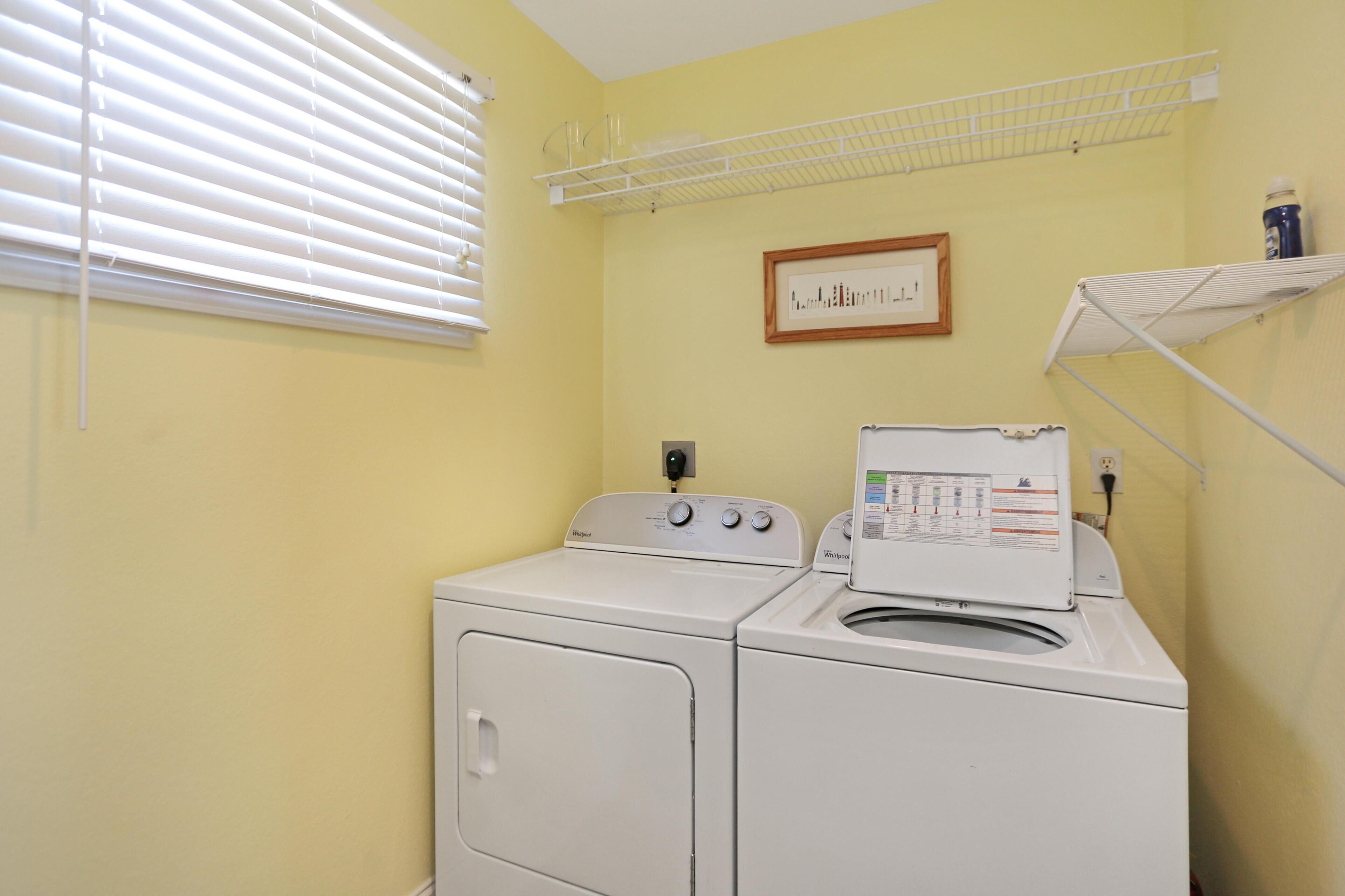 Une buanderie équipée de deux machines à laver et de deux sèche-linge blancs, d’une fenêtre avec des stores, d’une étagère sur laquelle est posée une photo encadrée et d’une prise de courant.