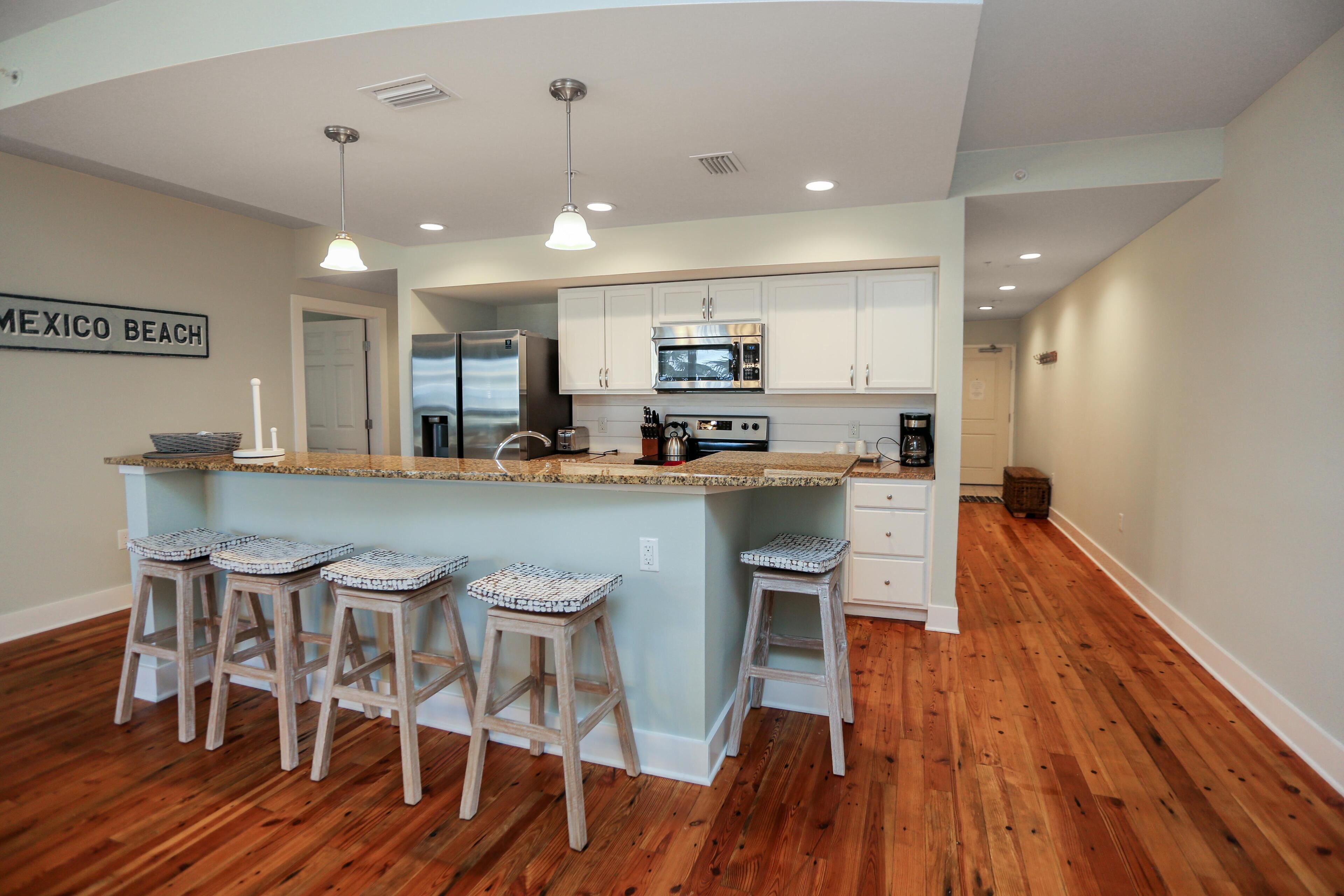 A modern kitchen with a central island, bar stools, white cabinetry, and a sign reading "MEXICO BEACH".