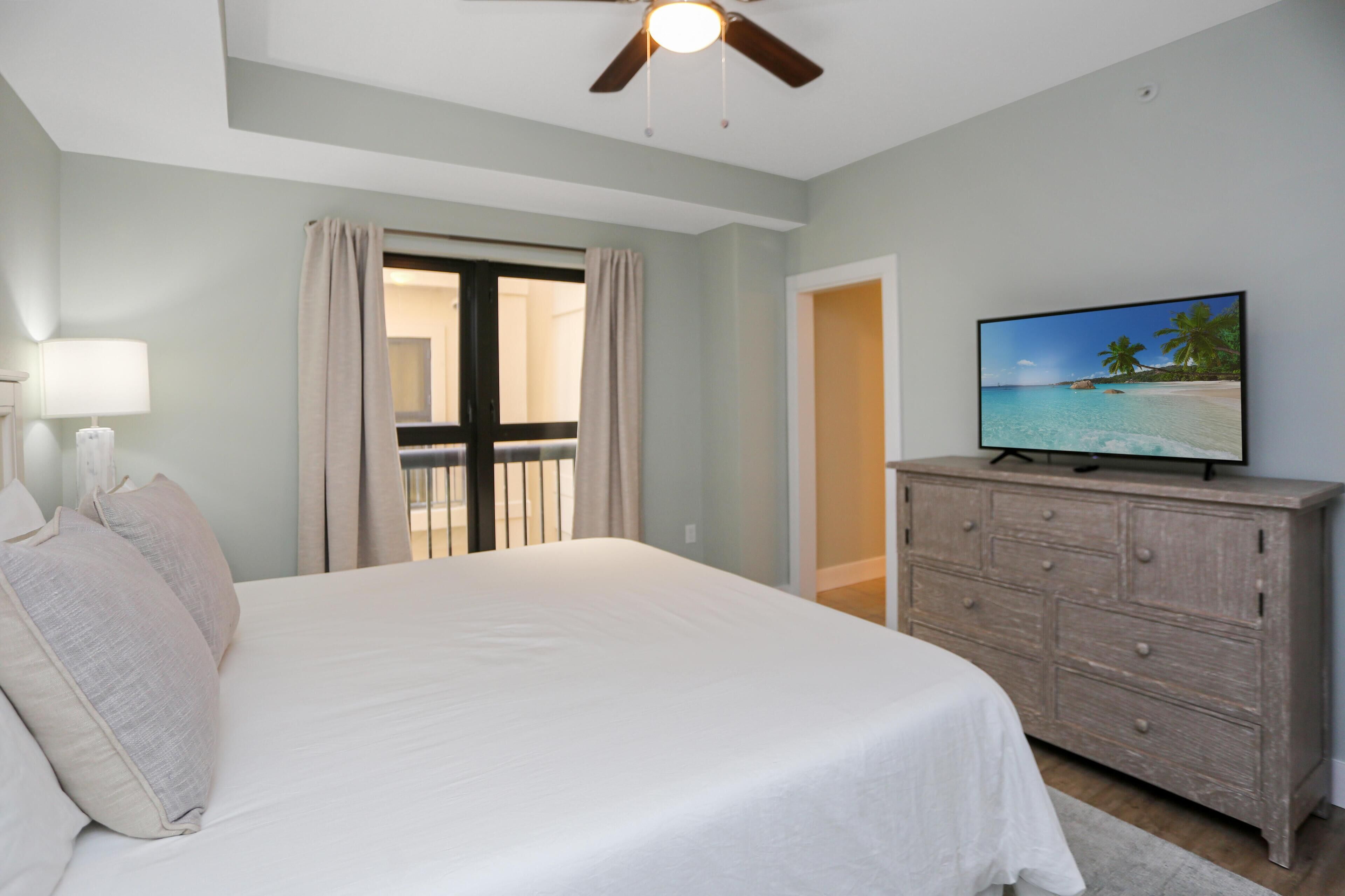 Une chambre à coucher avec un lit, une commode, un ventilateur de plafond et un téléviseur diffusant une scène de plage.