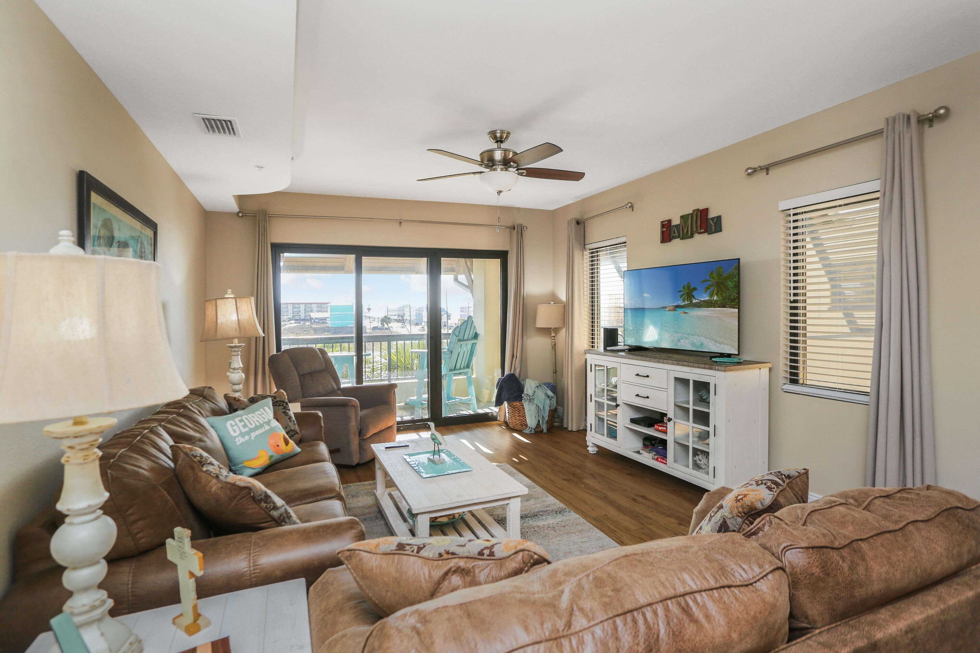 A living room with a brown leather sofa, a white coffee table, a television on a white cabinet, and a balcony with a view.