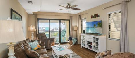 A living room with a brown leather sofa, a white coffee table, a television on a white cabinet, and a balcony with a view.