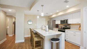 A modern kitchen with white cabinetry, a central island with bar stools, a stainless steel trash can, and a microwave above the oven. - Club at Mexico Beach 2D (Port St. Joe)