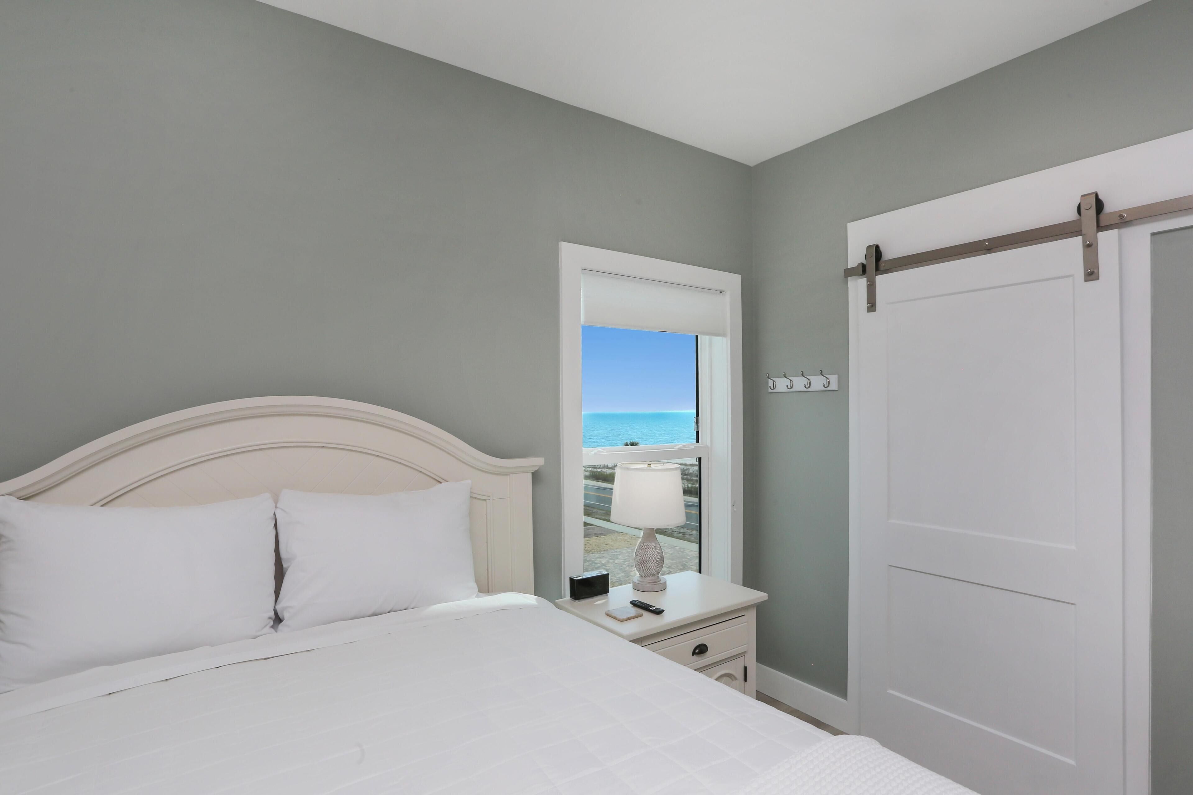 Une chambre à coucher avec un lit, une table de chevet et une vue sur la plage, visible par la fenêtre.