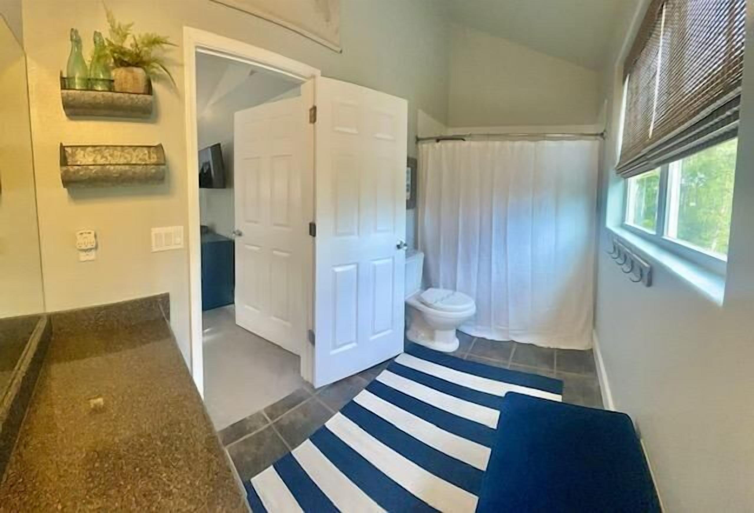 A bathroom with a white toilet, a shower curtain, a blue and white striped rug, and a door leading to another room.