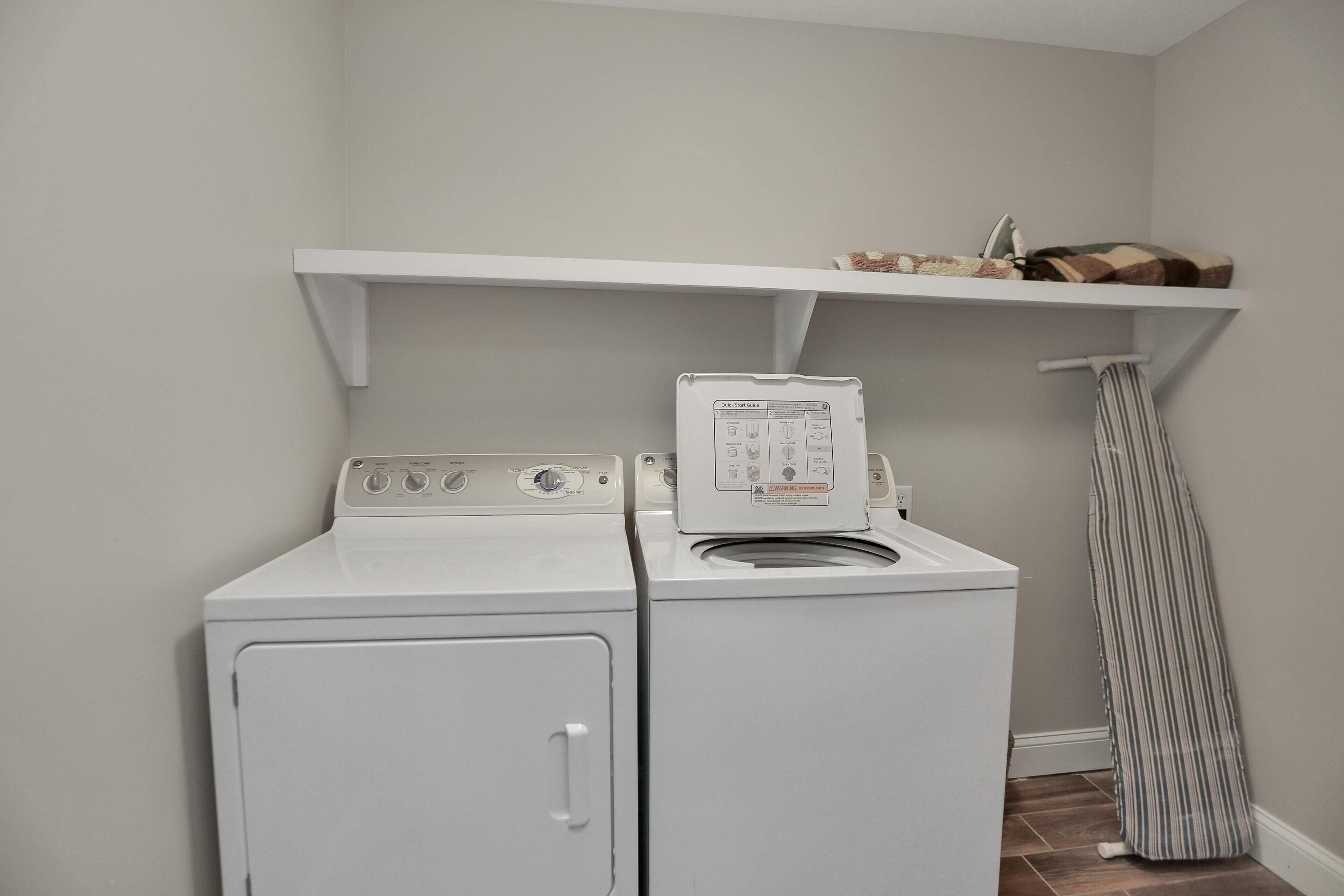 A laundry area with two white washing machines, a drying rack, and a shelf above the machines.