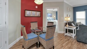 Een eetruimte met rieten stoelen en een glazen tafel, een woonkamer met een bank en een lamp, en een slaapkamer die door een open deur zichtbaar is.