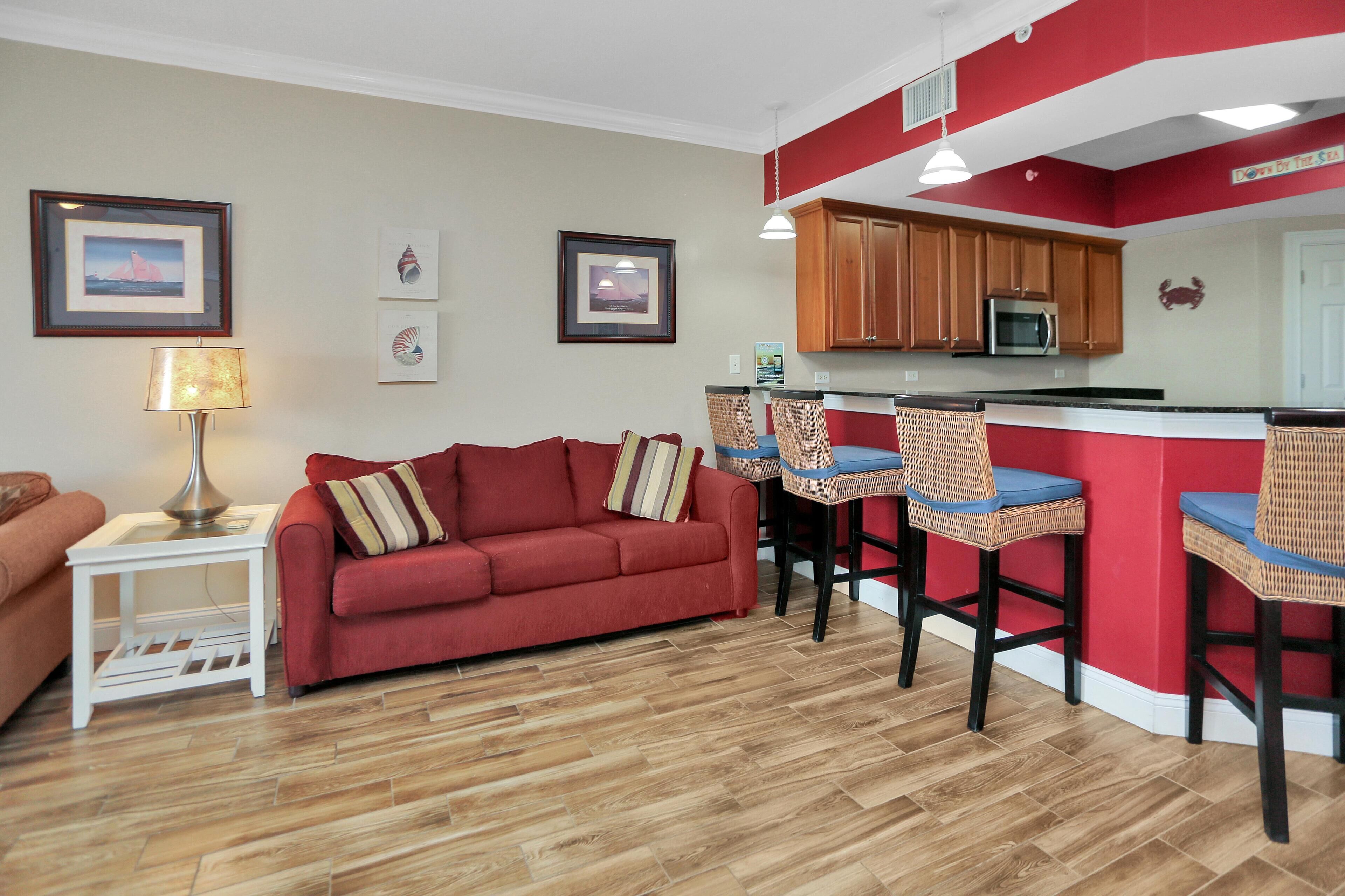 A compact living space with a red sofa, a small white side table, a lamp, a kitchen area with a microwave, and a bar counter with chairs.