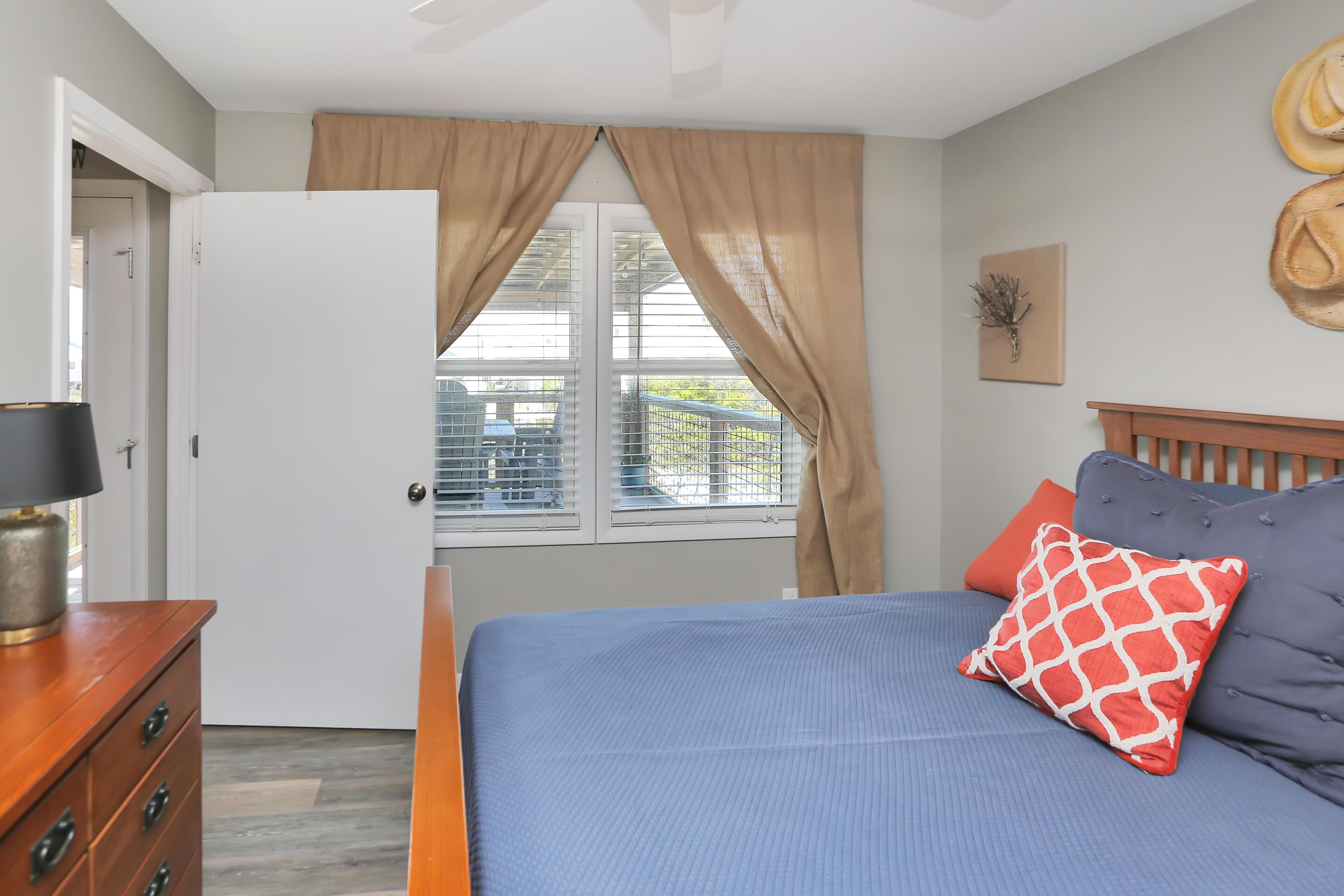 A bedroom with a bed, a wooden dresser, a window with curtains, and a view of the outside.