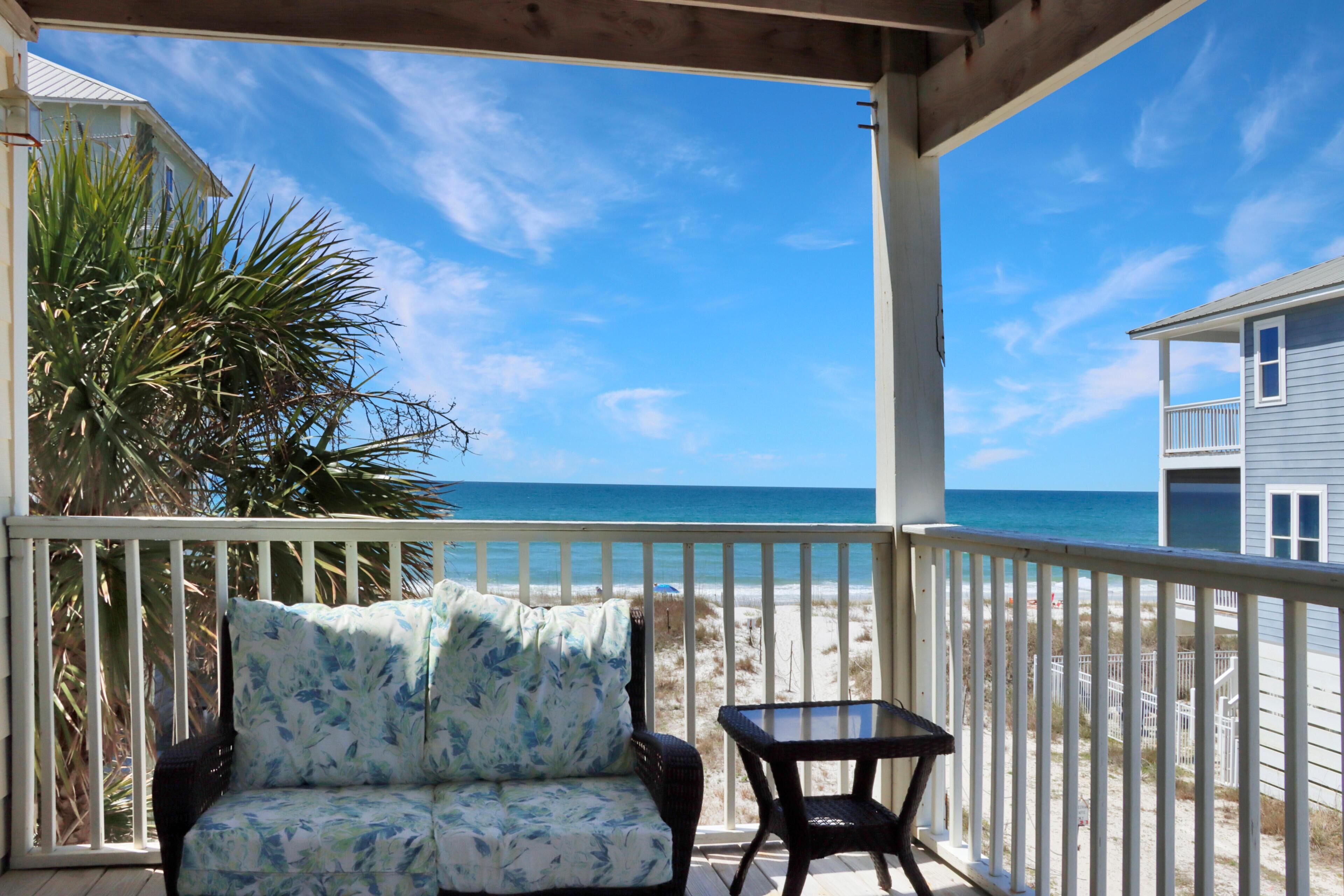 A covered outdoor area with a sofa and table, overlooking a beach and a building.