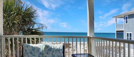 A covered outdoor area with a sofa and table, overlooking a beach and a building.