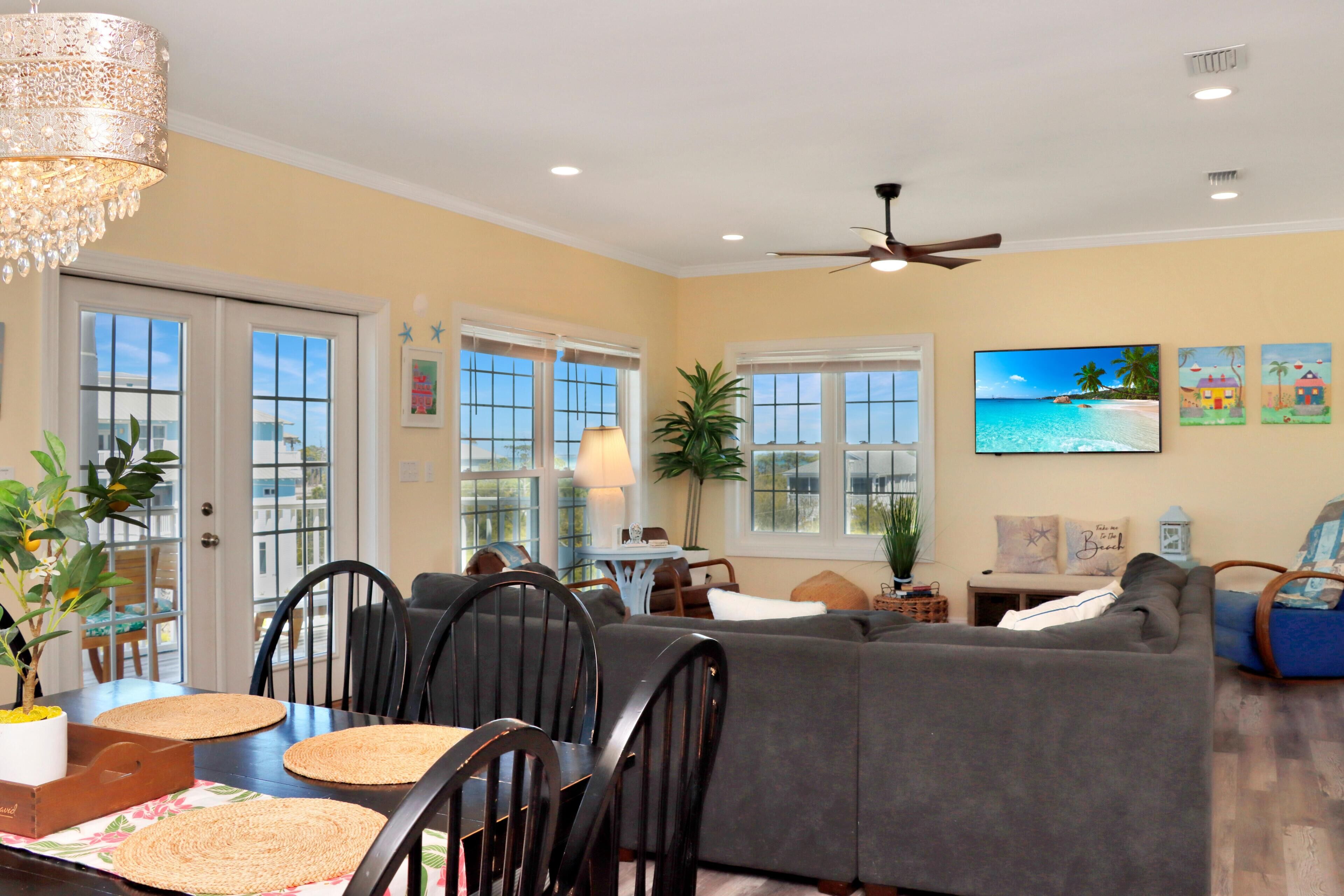 A living room with a sofa, dining table, chairs, a ceiling fan, and a TV on the wall.