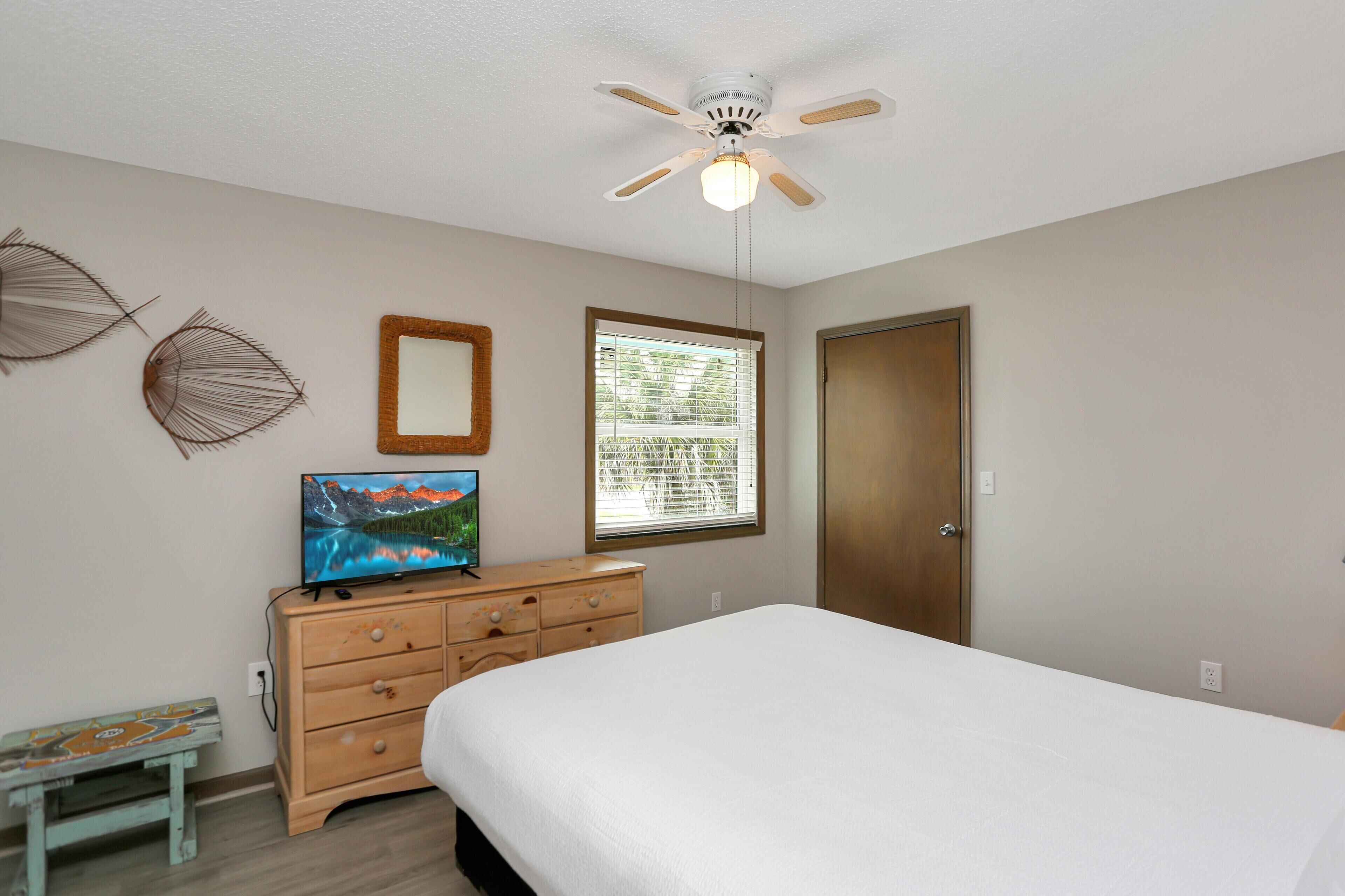 A bedroom with a bed, a dresser, a television, a ceiling fan, a window, and wall decorations.