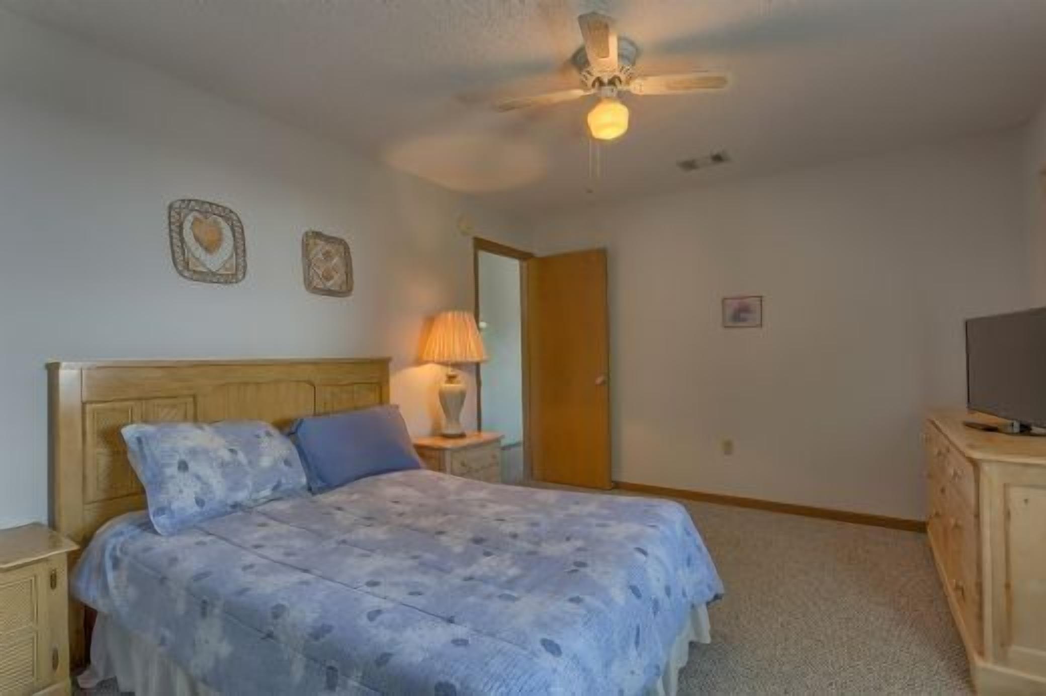 A bedroom with a bed, a ceiling fan, a nightstand, a lamp, a dresser, and a television.