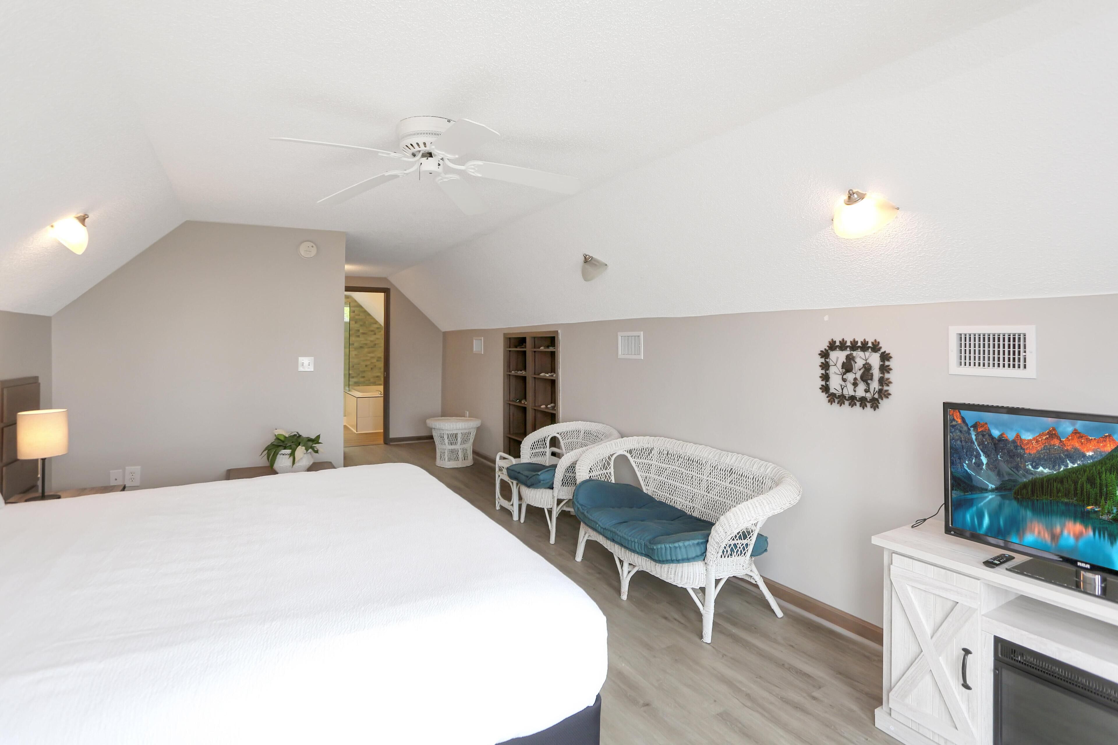 A spacious bedroom with a large bed, white wicker furniture, a television, and a ceiling fan.