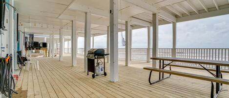 Una terraza cubierta de madera con un carrito para parrilla, bancos y vista al mar.