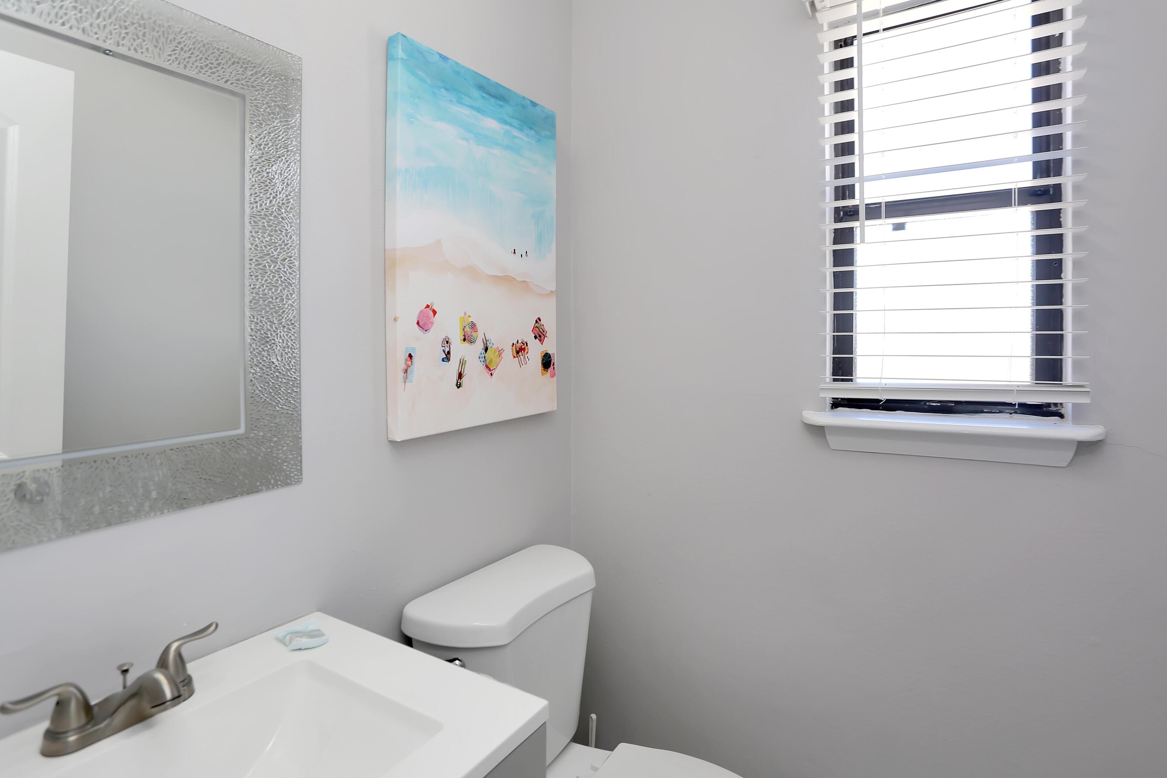 A bathroom with a white sink, a toilet, a mirror with a textured frame, and a window with blinds.