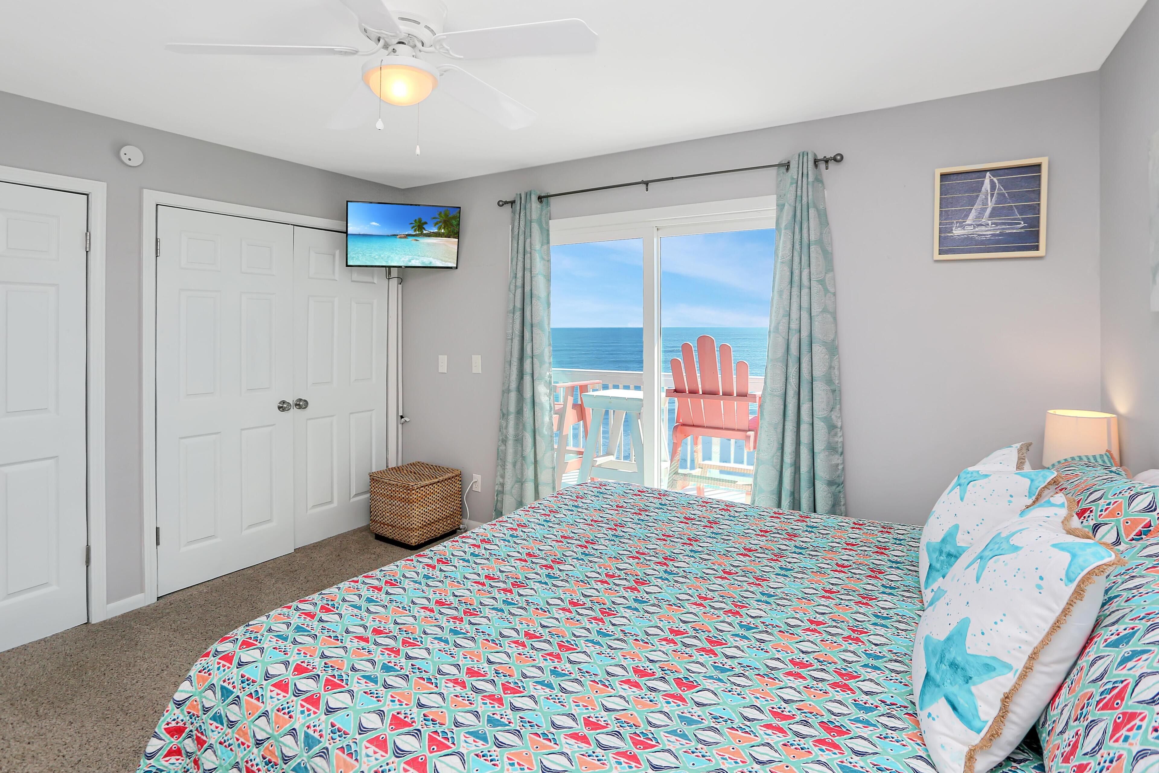 Une chambre à coucher comprenant un lit, un téléviseur, un ventilateur de plafond et une vue sur l’océan, accessible par une porte coulissante.