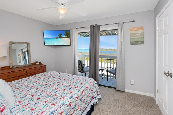 A bedroom with a bed, a ceiling fan, a TV, a dresser, and a balcony with a view of the ocean.
