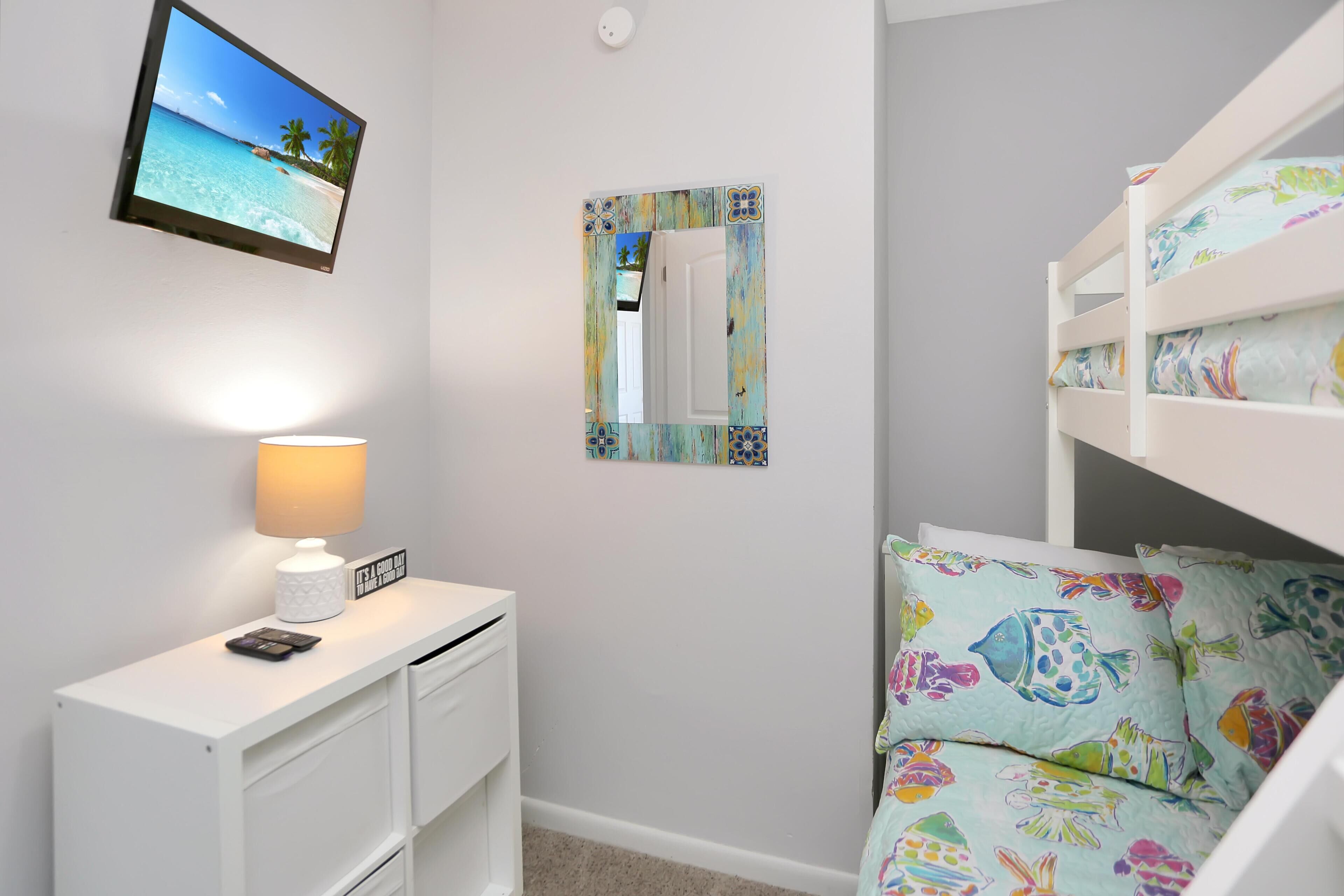 Une chambre avec un lit superposé, une table de chevet avec une lampe, un petit bureau et un téléviseur fixé au mur qui diffuse une scène de plage.