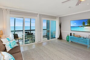 A living room with a view of the ocean, a sliding glass door, a sofa with decorative pillows, a television, and a blue console table. - The Hidden Pearl (Port St. Joe)