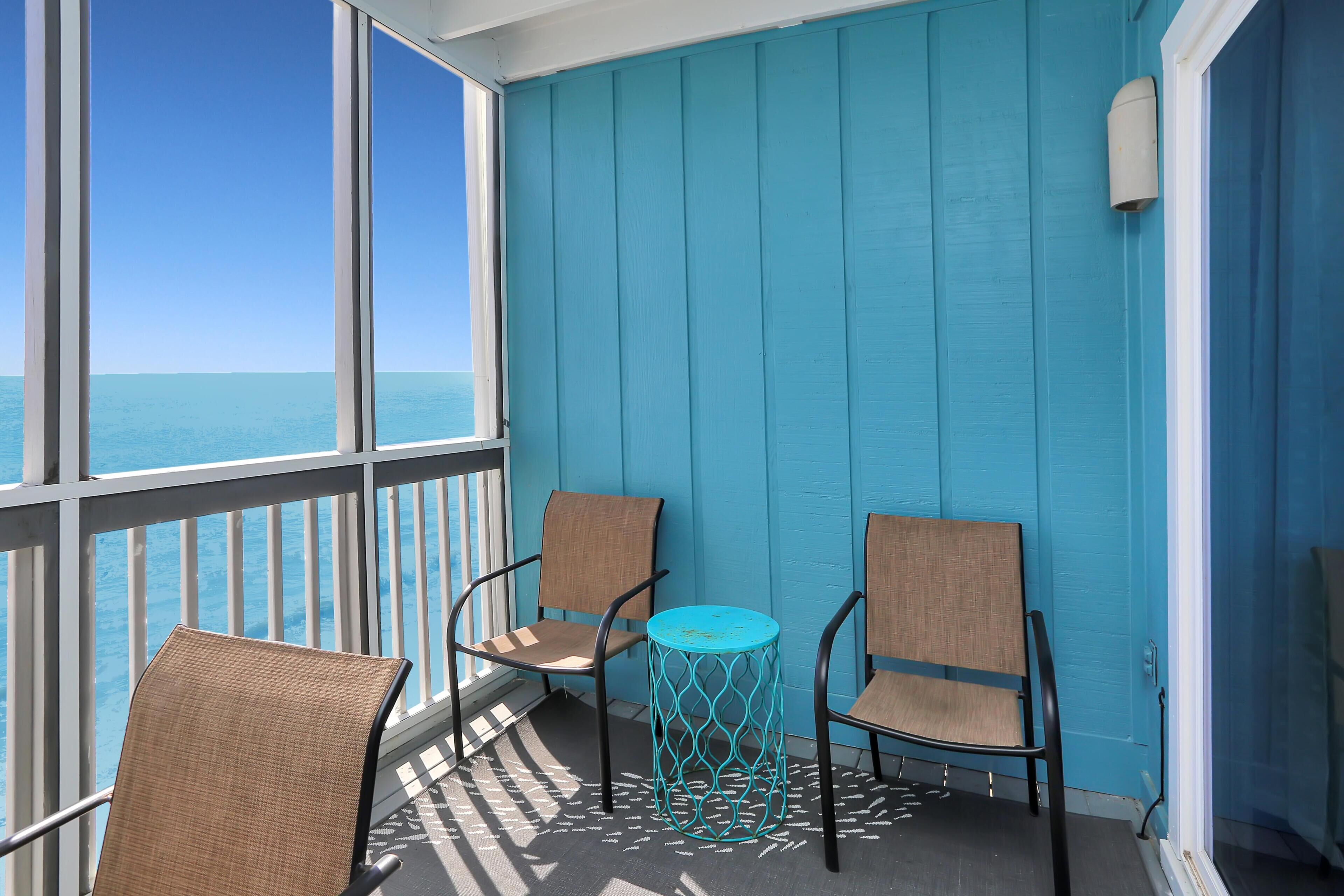 A balcony with two wicker chairs and a small turquoise table, overlooking a body of water.