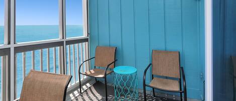 A balcony with two wicker chairs and a small turquoise table, overlooking a body of water.