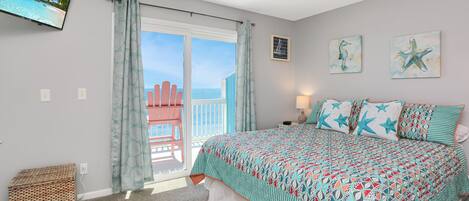 A bedroom with a bed, a television on the wall, and a balcony with a view of the ocean.