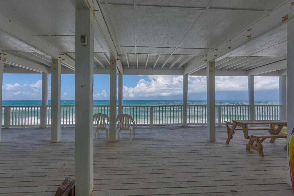 An open-air deck with white railings, wooden benches, and chairs facing a beach.